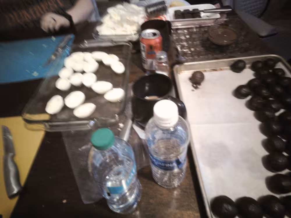 Image of a cluttered table where people are preparing food. On the left are cutting boards and hard boiled eggs getting cut in half to be made into deviled eggs. On the right are bite-sized balls of oreos about to be coated in melted chocolate. Admist this are bottles of water, cans of soda, and mugs of hot chocolate.